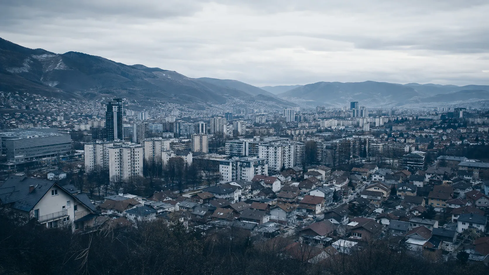A city in Bosnia and Herzegovina from a hillside on an overcast day, blocks and houses along a valley under mountains