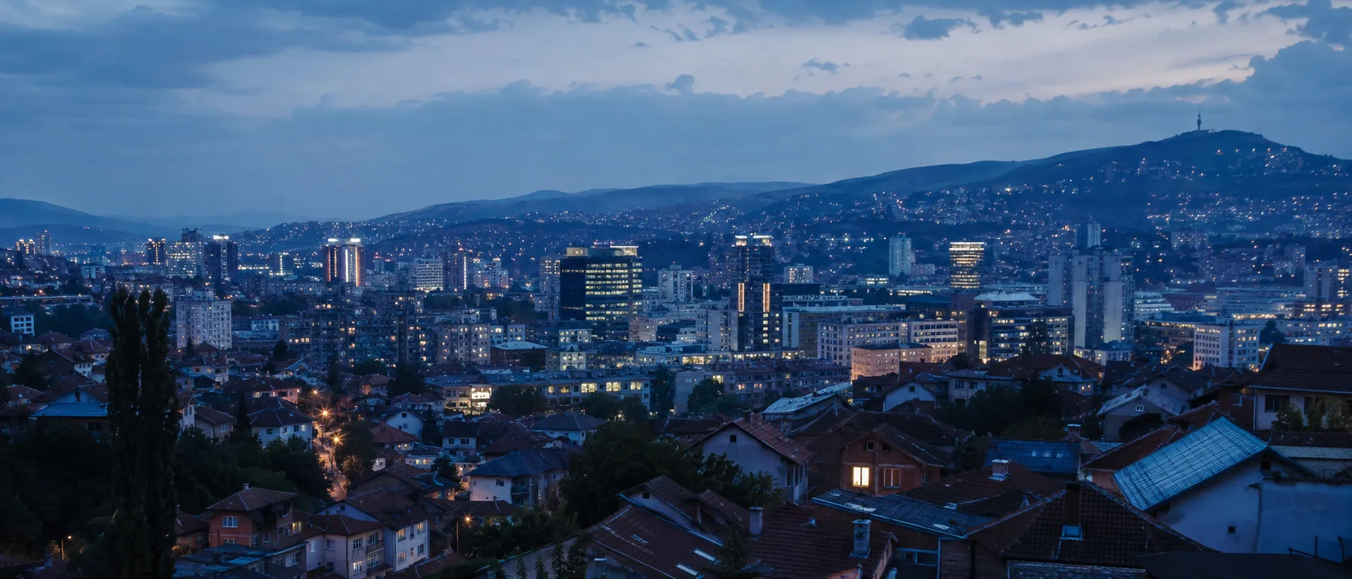 A city in Bosnia and Herzegovina at dusk, mid-rise blocks and tiled rooftops below a mountain ridge